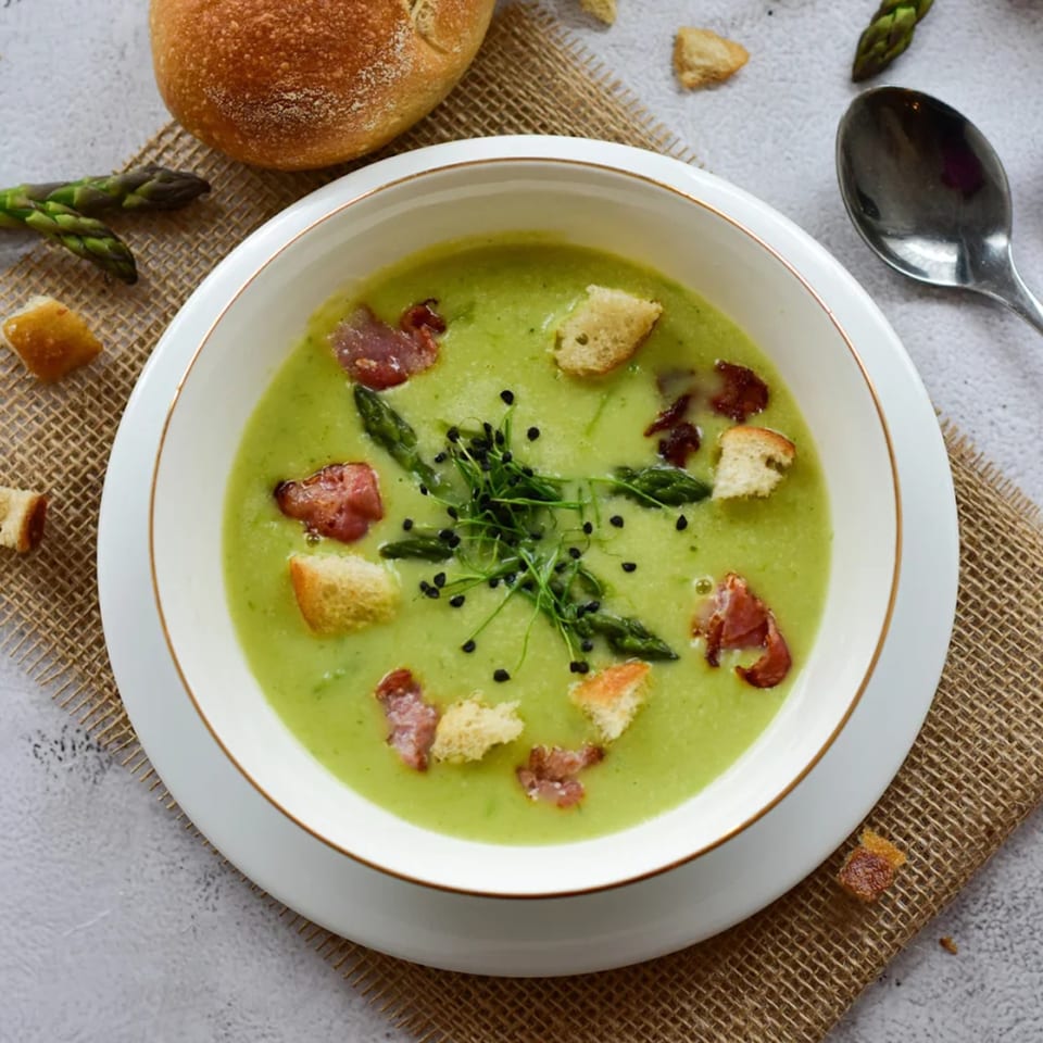 Creamy green soup with asparagus, bacon, croutons, and microgreens, served with a bread roll.
