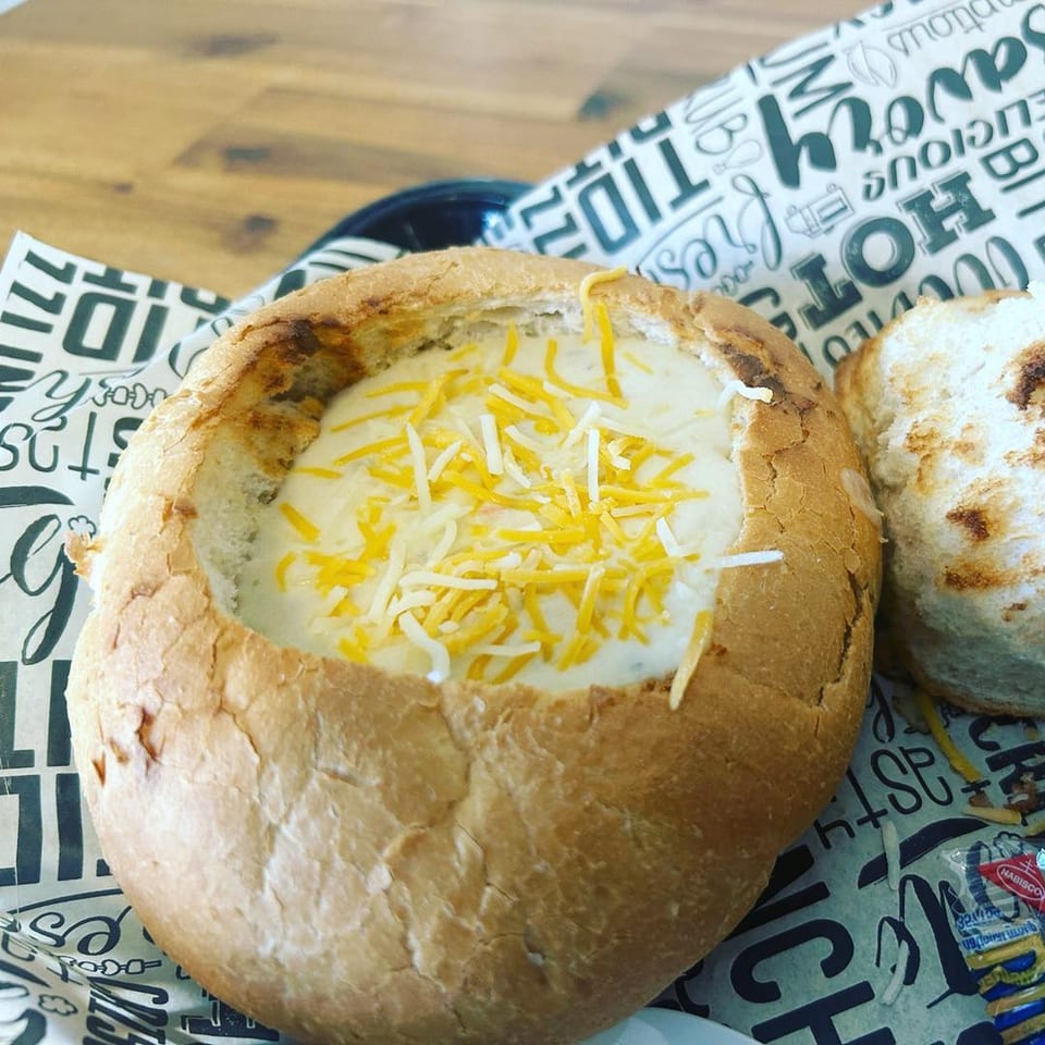 A bread bowl filled with creamy soup topped with shredded cheese, served with a side of toasted bread.