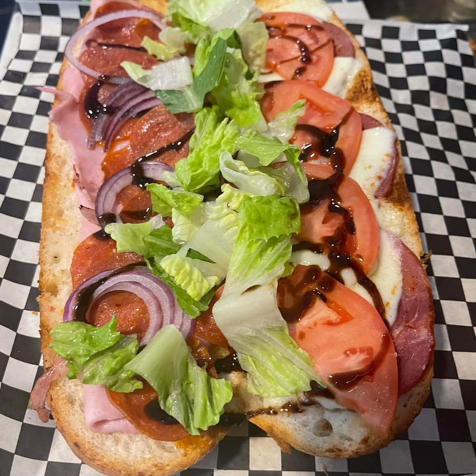 A Grinders Soups and Subs sandwich with meats, cheese, tomatoes, red onion, lettuce, and balsamic glaze.
