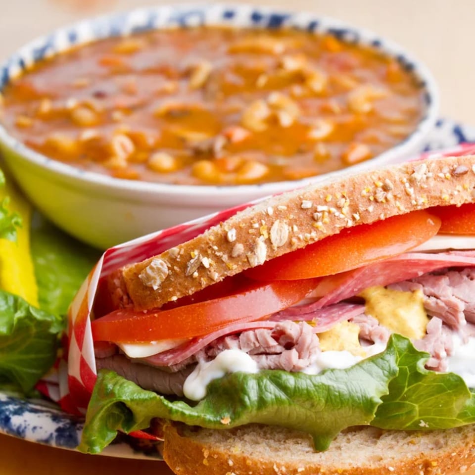A Grinders Soups and Subs sandwich with meat, cheese, lettuce, tomato, and sauces, next to a bowl of soup.