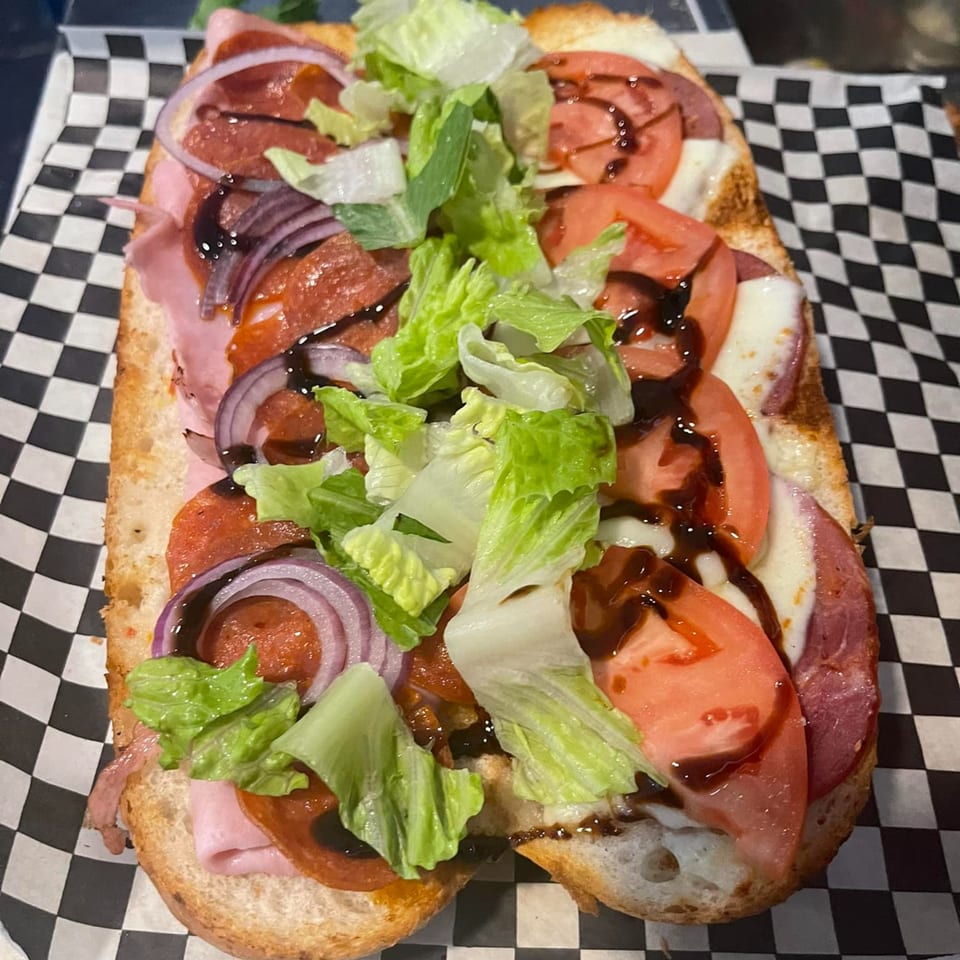 A Grinders Soups and Subs sandwich with meats, cheese, tomatoes, onions, lettuce, and balsamic glaze.