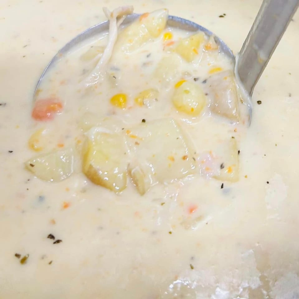 A ladle scoops creamy potato soup with corn, carrots, and mushrooms from a large pot.