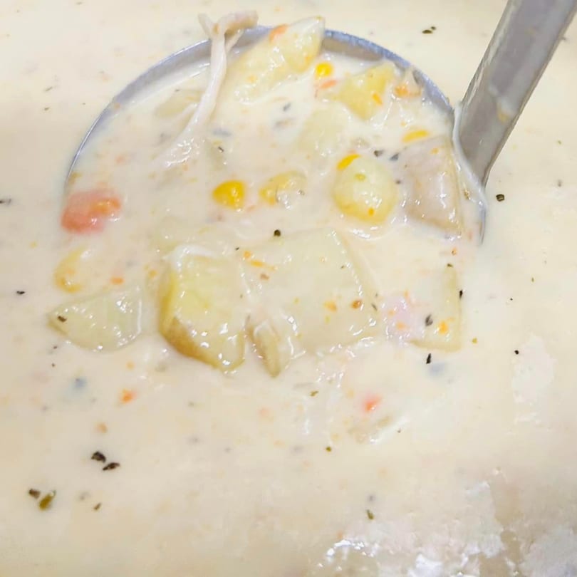A ladle scoops creamy potato soup with corn, carrots, and mushrooms from a large pot.